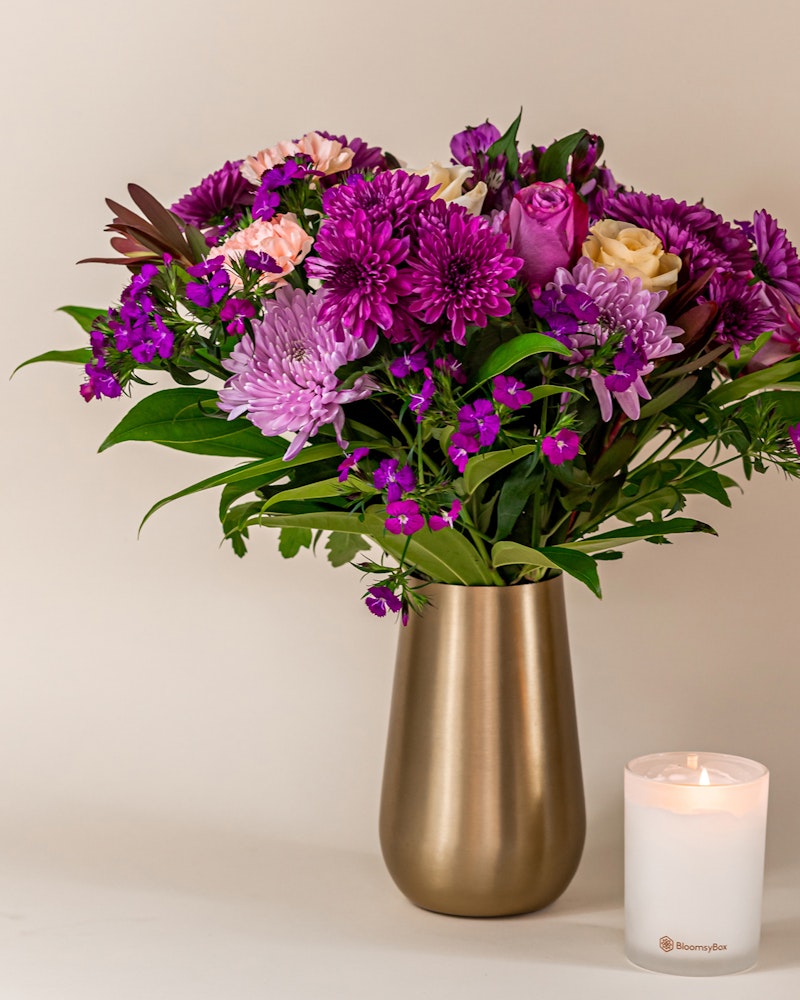 A vibrant bouquet of purple and pink flowers with touches of white in a gold vase, accompanied by a lit white candle on a neutral background.