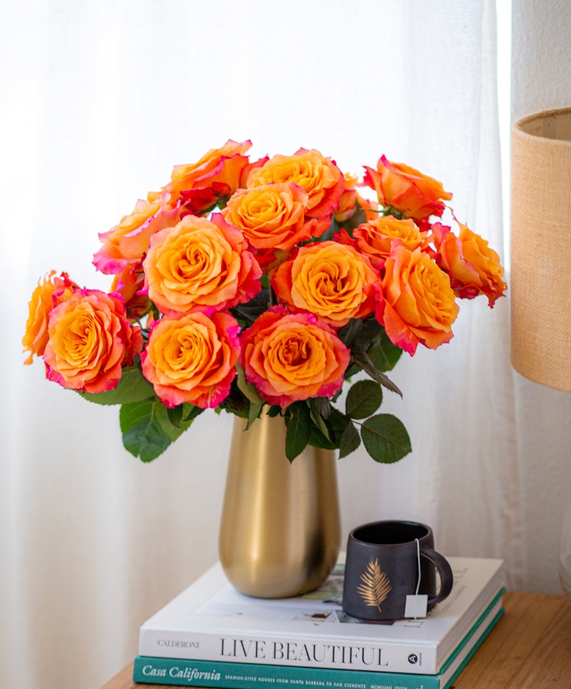 A vibrant bouquet of orange roses with pink tips arranged in a golden vase on a stack of books beside a black mug, lit by natural light near a window with sheer curtains.