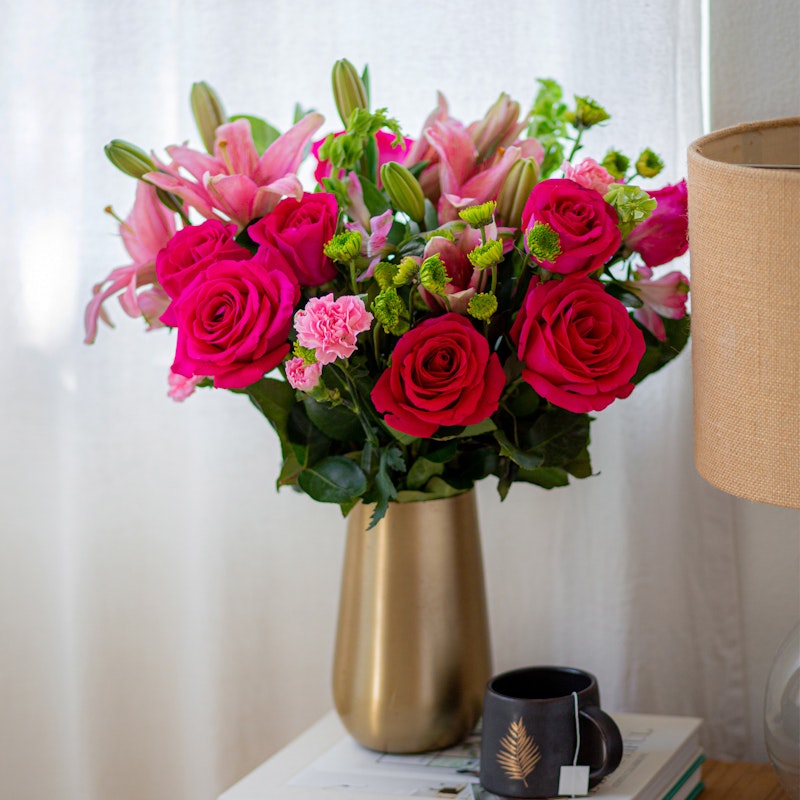 Bouquet of vivid roses and lilies in a golden vase on a white table with a coffee mug, books, and a lamp, beside a window with white curtains.