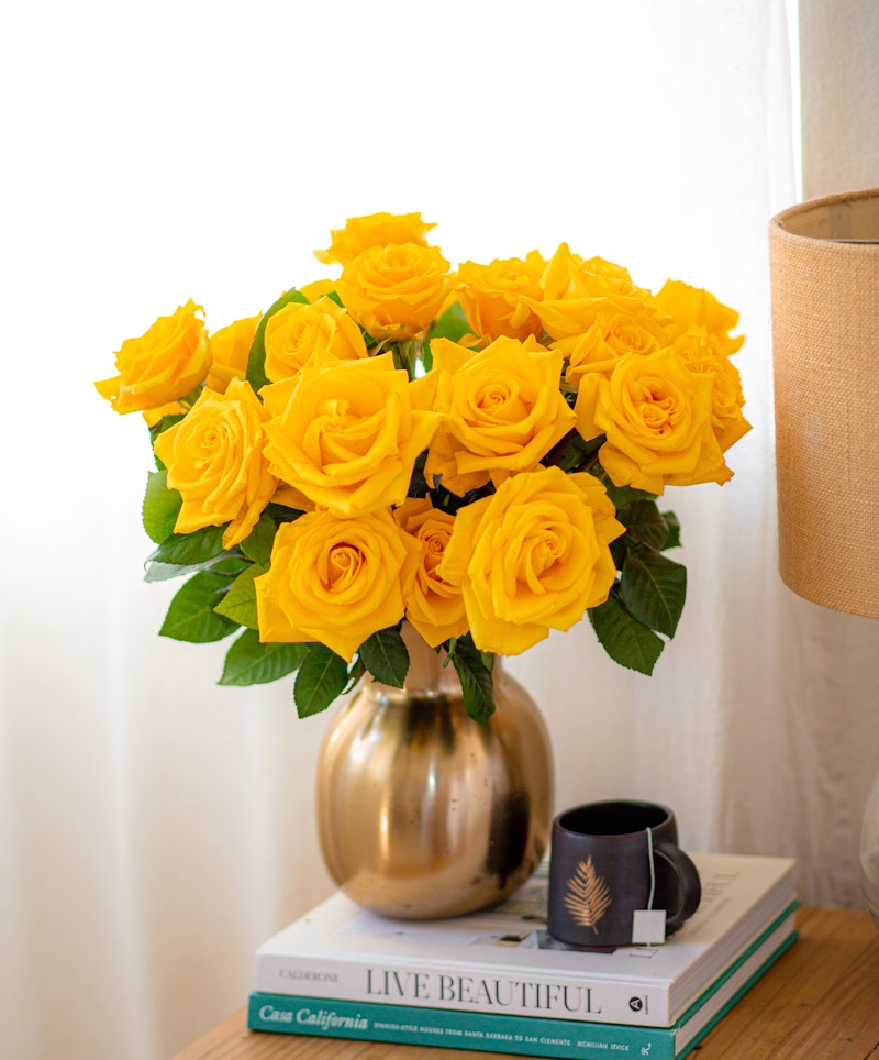 A vibrant bouquet of fresh yellow roses arranged in a gold vase, placed on top of two decorative books beside a small black cup on a wooden table.