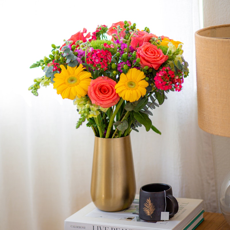 Bright and colorful bouquet of flowers including yellow gerberas and pink roses in a gold vase on a table beside a small black cup and books.