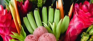Vibrant tropical fruit and flower arrangement featuring ripe green bananas, red ginger, and bird of paradise flowers, with subtle pink textures added by loofah sponges.