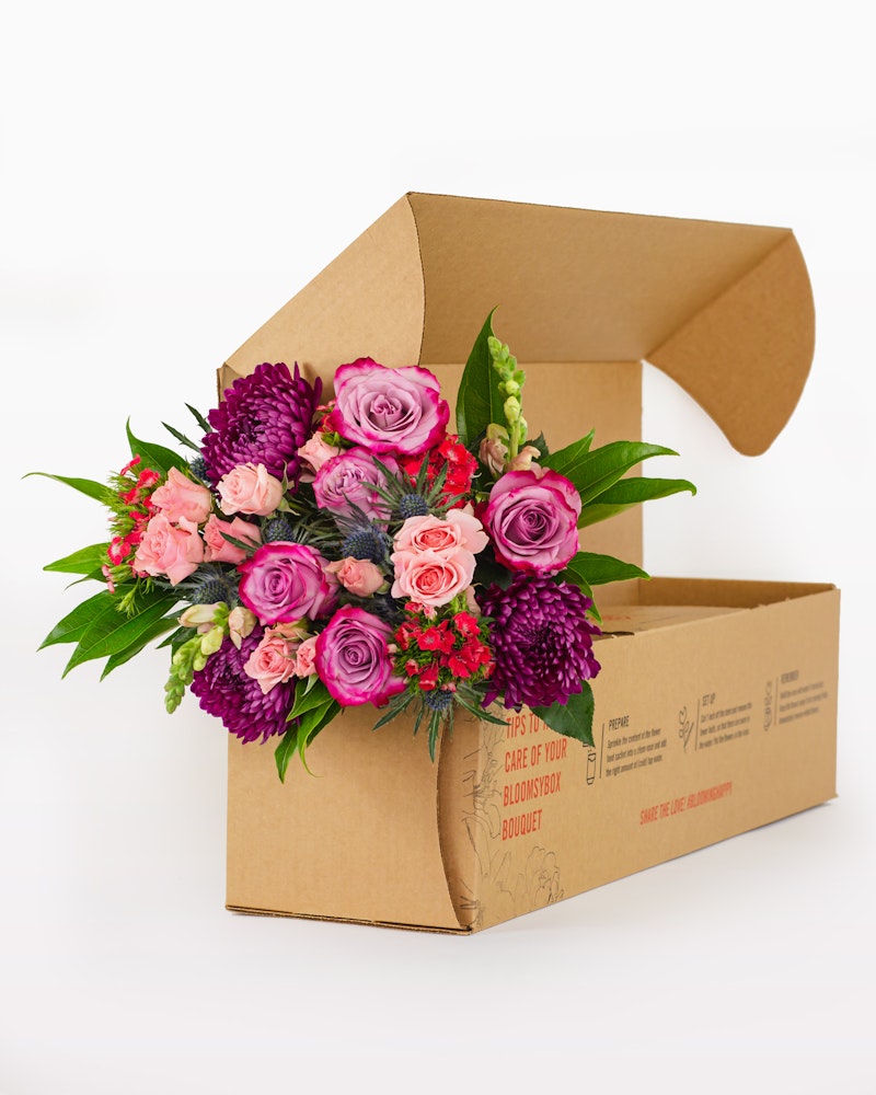 Vibrant bouquet of fresh flowers with pink roses and purple accents emerging from a cardboard flower box against a white background, ready for delivery.