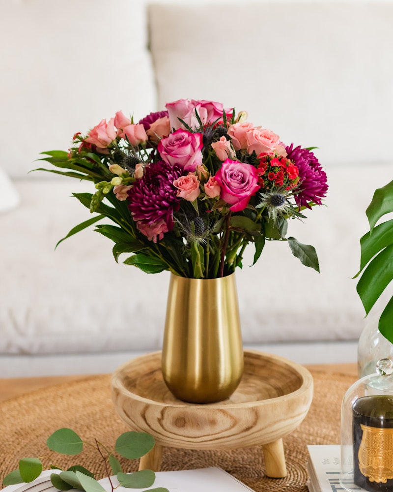 A vibrant bouquet of pink roses and assorted flowers in a gold vase on a wooden tray, with a cozy white couch and green plant leaves in the background.