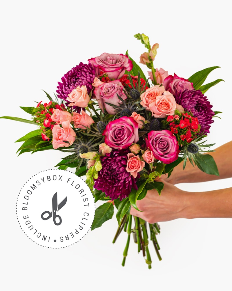 A beautiful bouquet of pink and purple flowers, including roses and chrysanthemums, held by a person against a white background, showcased by BloomsyBox Florist Selection.