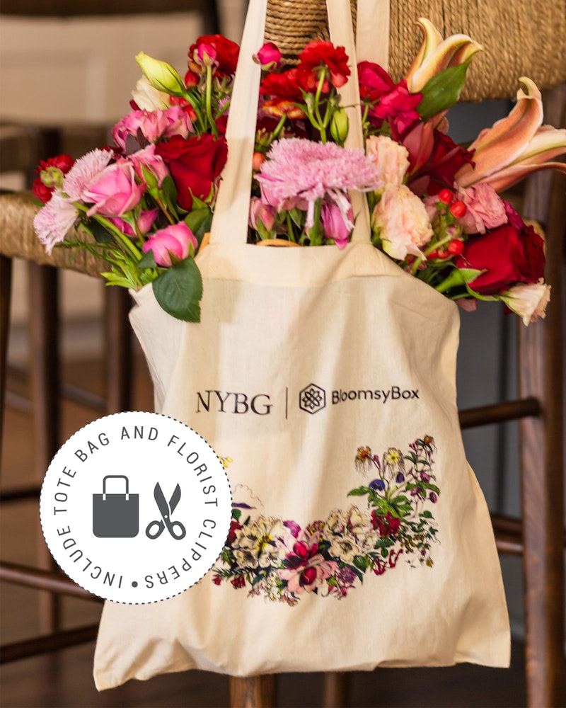 A canvas tote bag from NYBG by BloomsyBox, adorned with a floral print design, hangs on a chair filled with a fresh bouquet of red, pink, and purple flowers.