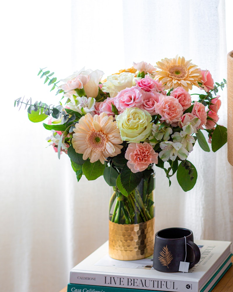 A vibrant bouquet of pink roses, gerberas, and assorted greenery in a glass vase on a table with books and a black coffee mug, beside a window with sheer curtains.