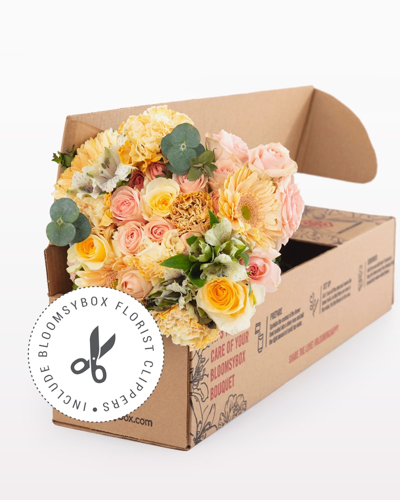 Bouquet of fresh flowers with roses and chrysanthemums in shades of yellow and pink, elegantly arranged and packaged in a BloomsyBox branded cardboard box.