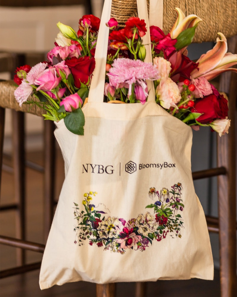 A colorful bouquet of fresh flowers, including roses and lilies, overflows from a beige BloomsyBox tote bag with floral print hanging on a wooden chair's back.