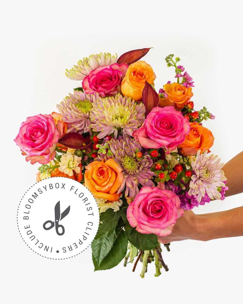 Vibrant bouquet of fresh flowers including pink roses, purple chrysanthemums, and orange accents against a white background, with a 'BloomsBox Florist Service' logo.