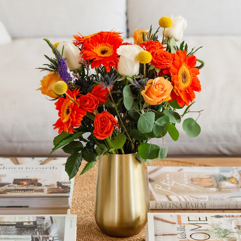 Colorful bouquet of flowers including orange gerberas, red roses, and white tulips in a golden vase on a coffee table with assorted books.