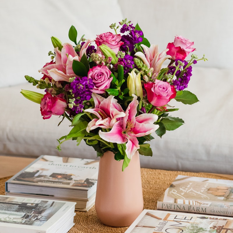 Elegant floral arrangement in a blush vase with pink lilies, purple flowers, and roses, displayed on a table with decorative books in a cozy home setting.