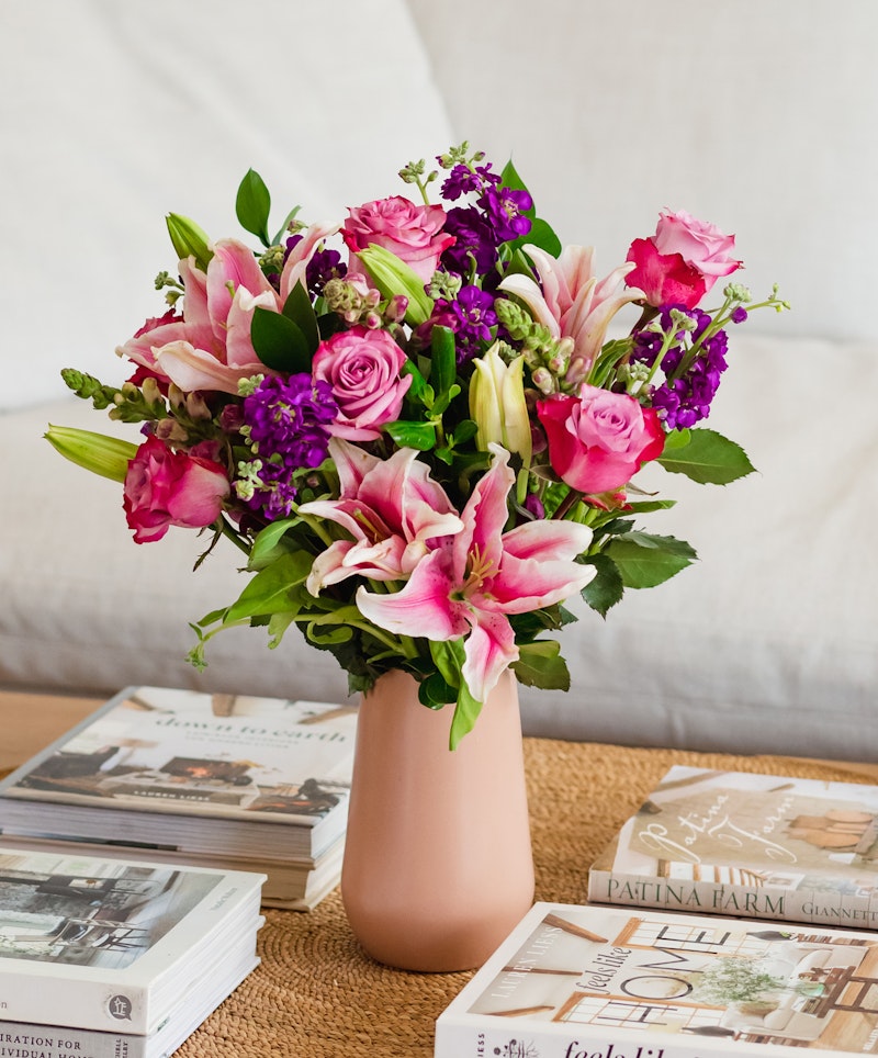 A vibrant bouquet of pink roses and lilies with purple accents in a pastel vase, set on a table with stylish books about home design and decoration.