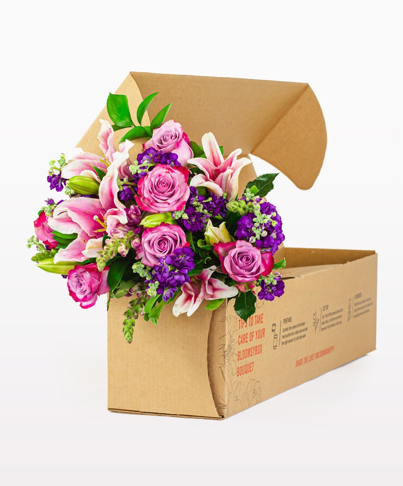 Vibrant bouquet of pink and purple flowers with lush green leaves elegantly arranged and partially nestled in an open brown cardboard box against a white background.