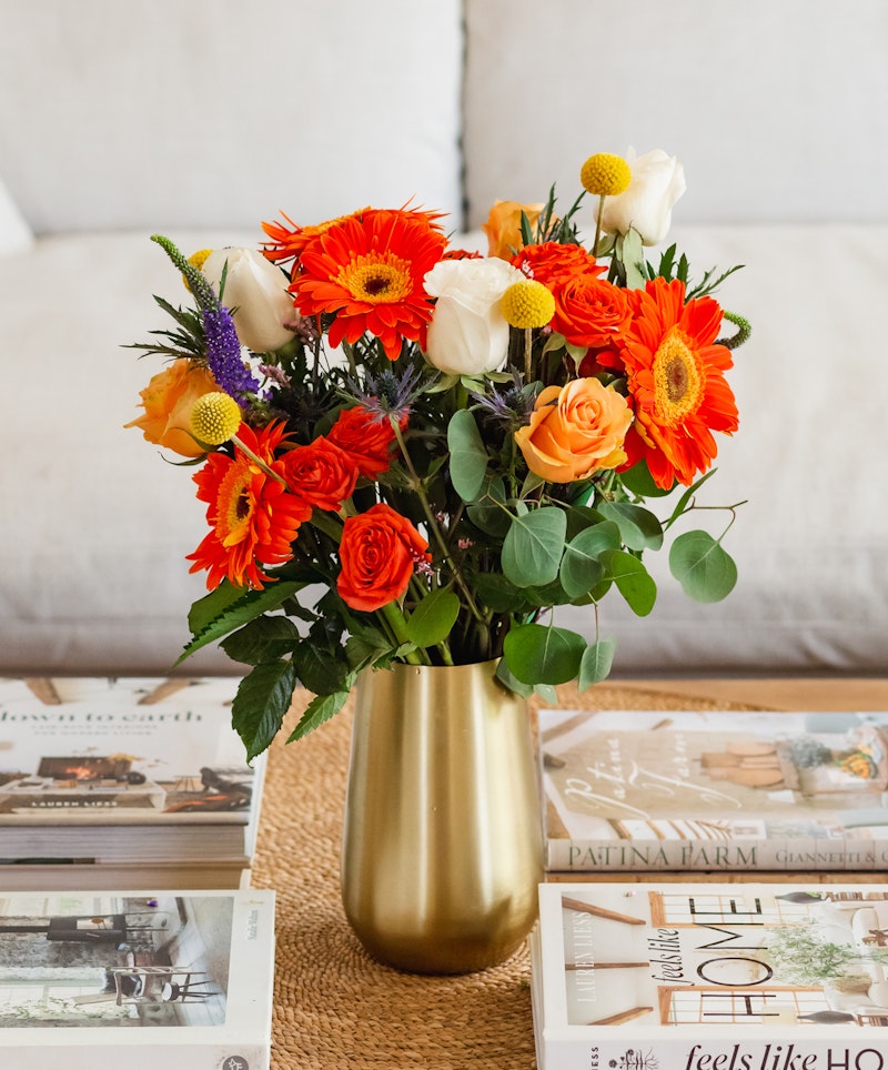 Vibrant bouquet of orange gerberas, roses, and white blooms in a gold vase on a table with decorative magazines, creating a cozy and elegant home setting.