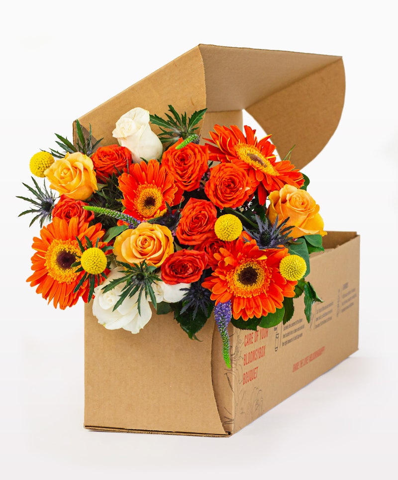 Vibrant bouquet of fresh flowers including orange gerberas, yellow sunflowers, and white roses spilling out of a brown cardboard box on a white background.
