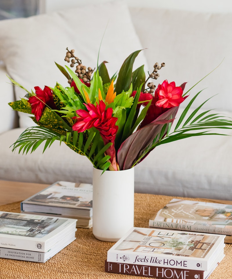 A vibrant bouquet of tropical flowers in a white vase sits on a wooden table surrounded by books on home and travel, adding a cozy and inviting atmosphere to the room.