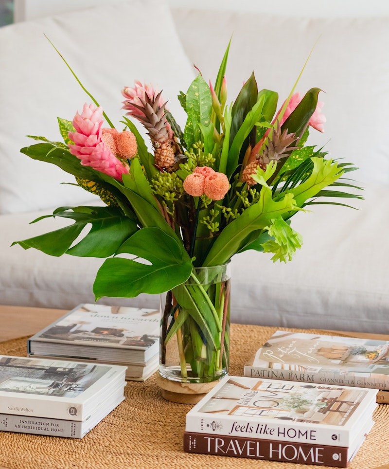 A vibrant bouquet of pink and orange flowers arranged in a clear glass vase on a wooden table, with decor magazines splayed out beneath, in a cozy home setting.