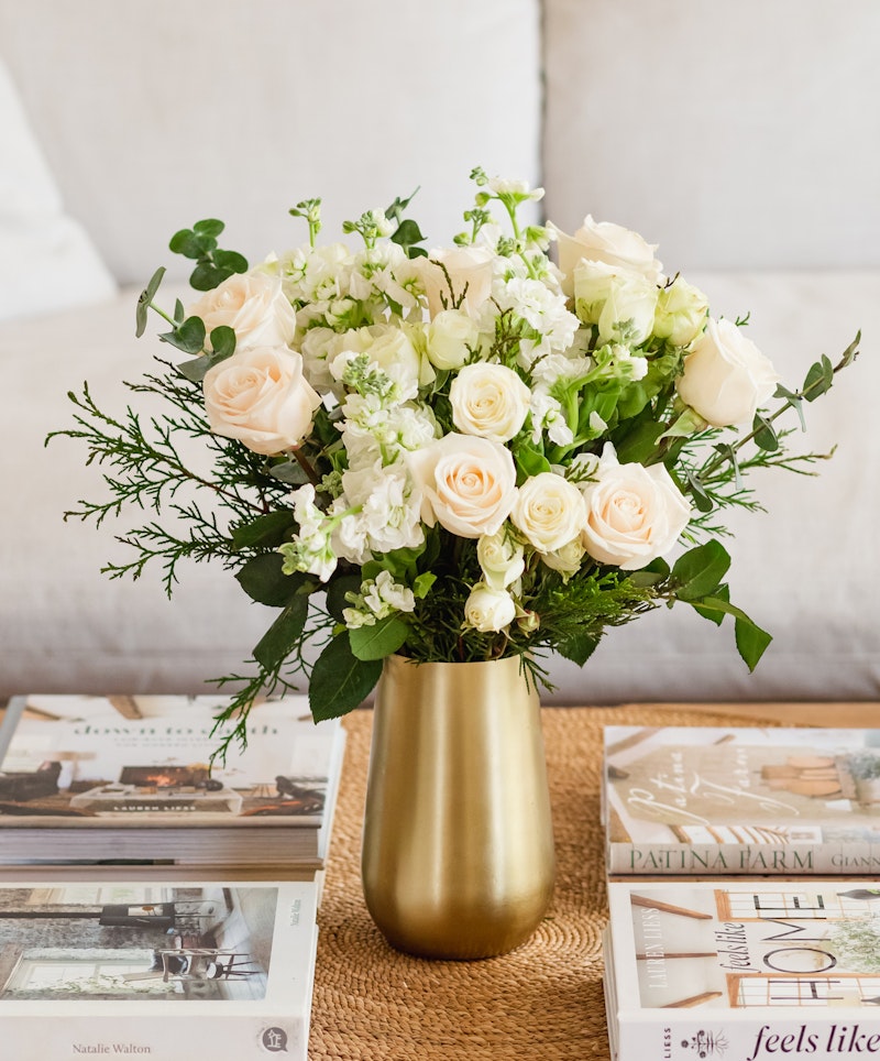 Elegant bouquet of pale pink roses and white flowers arranged in a gold vase on a table with assorted interior design books, creating a sophisticated home decor ambiance.