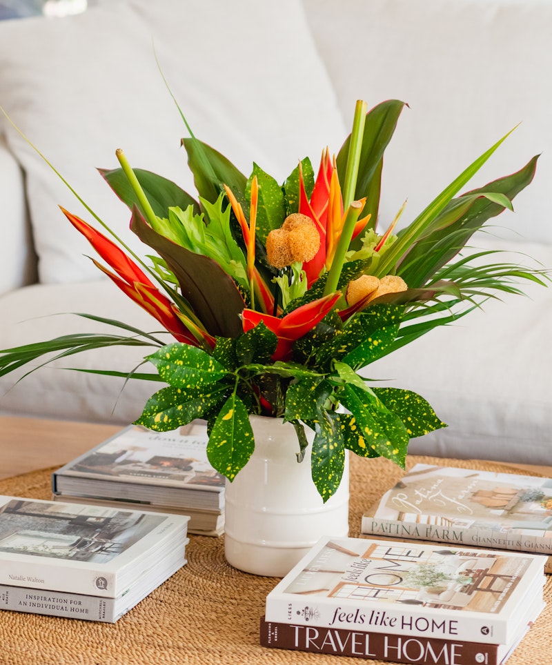 A vibrant bouquet of tropical flowers in a white vase sits atop a table alongside neatly stacked lifestyle books, with a cozy beige couch in the background.