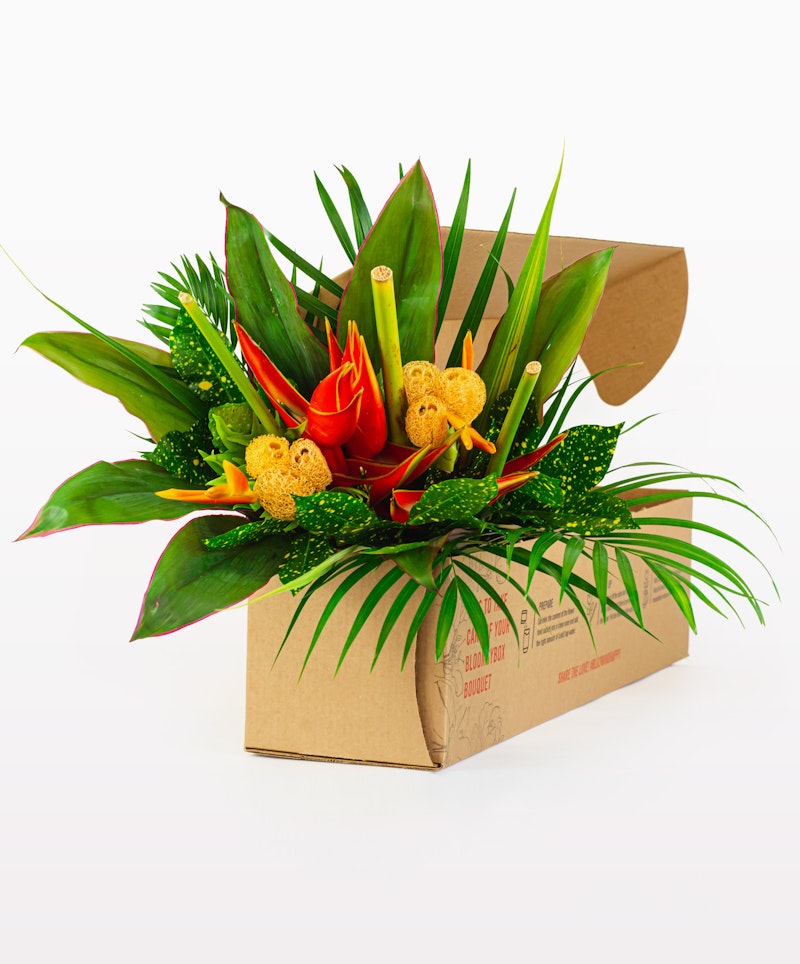 Vibrant tropical flower arrangement with red heliconia, yellow sponge mushrooms, and lush green leaves in a cardboard box against a white background.