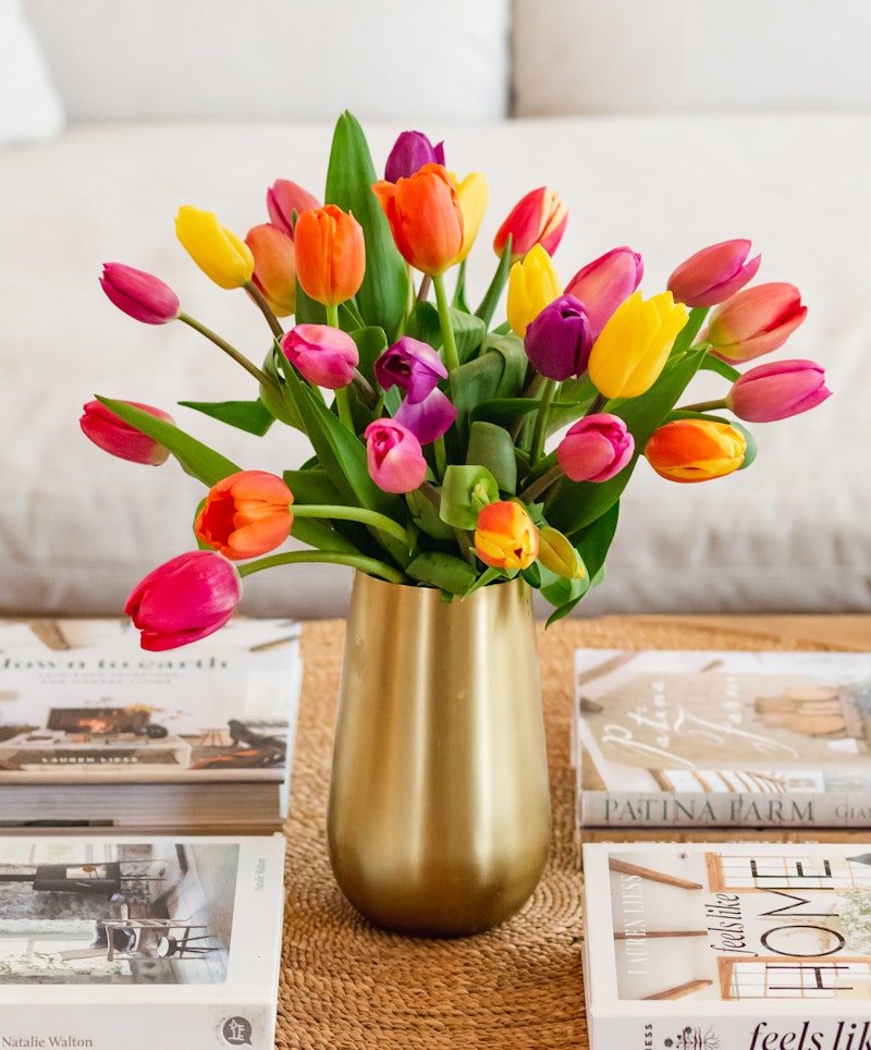 A vibrant bouquet of multicolored tulips arranged in a gold vase on a table with decorative books about home and garden in a cozy living room setting.
