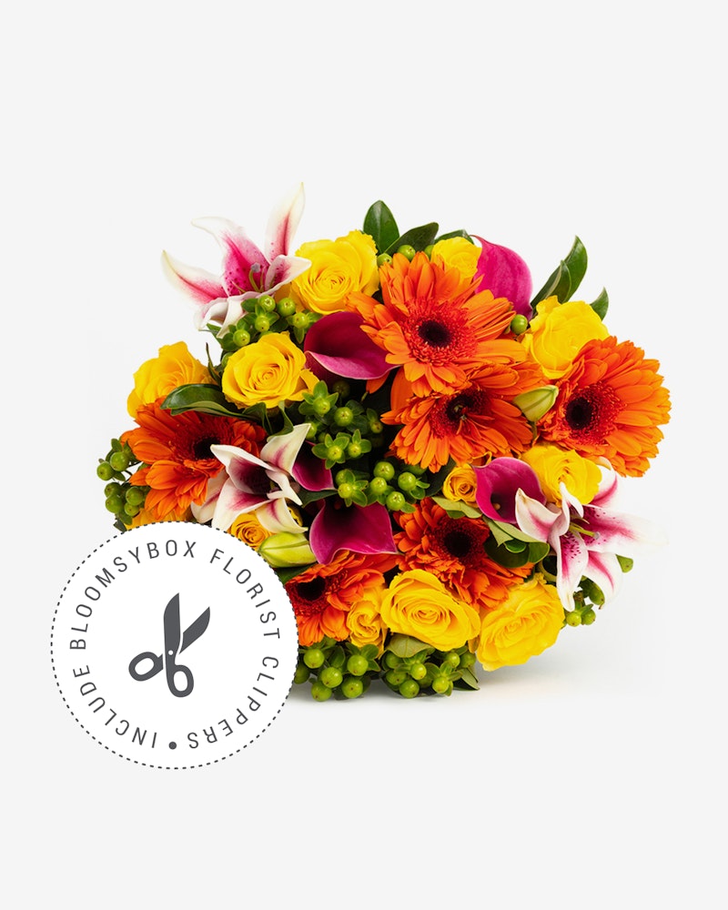 Vibrant bouquet of fresh flowers including orange gerberas, yellow roses, pink lilies, and green accents arranged by BloomsBox Florist on a white background.