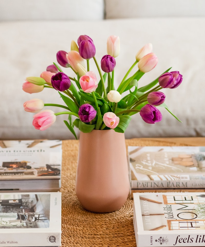 A vibrant bouquet of pink and purple tulips arranged in a modern pink vase, set on a woven mat over a coffee table with stylish home decor books.