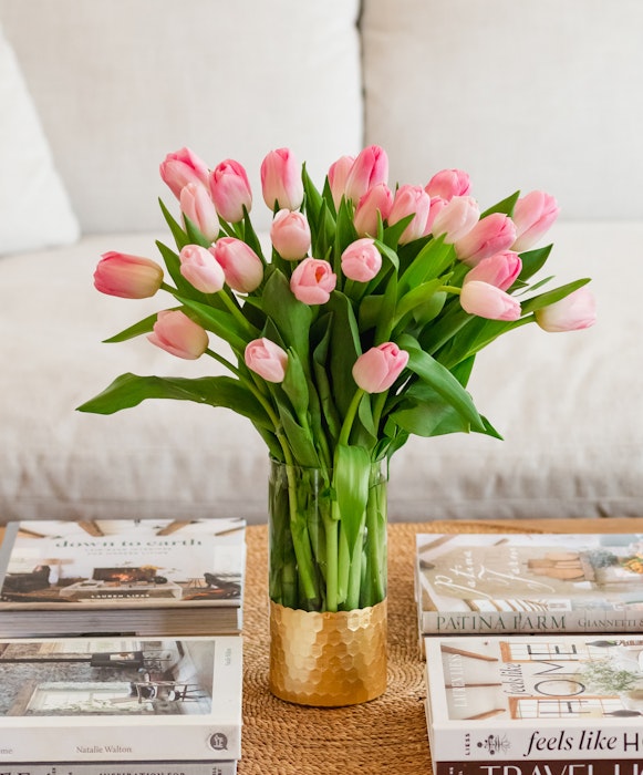 A vibrant bouquet of pink tulips elegantly arranged in a hammered gold vase on a coffee table, surrounded by an array of stylish interior design books.