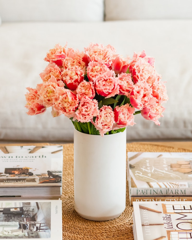 A bouquet of pink fringed tulips in a white vase on a woven mat, with lifestyle magazines scattered around on a wooden coffee table in a cozy living room setting.