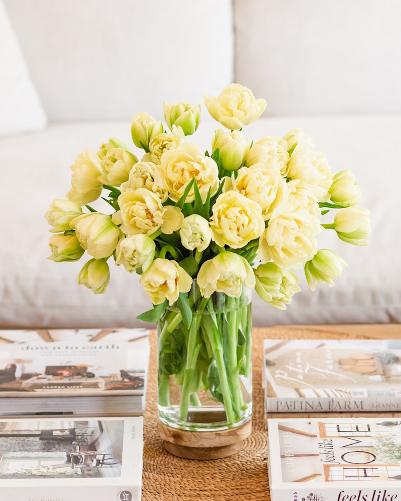 A vibrant bouquet of yellow tulips in a clear glass vase on a wooden coffee table, surrounded by assorted cozy living room decor and lifestyle magazines.