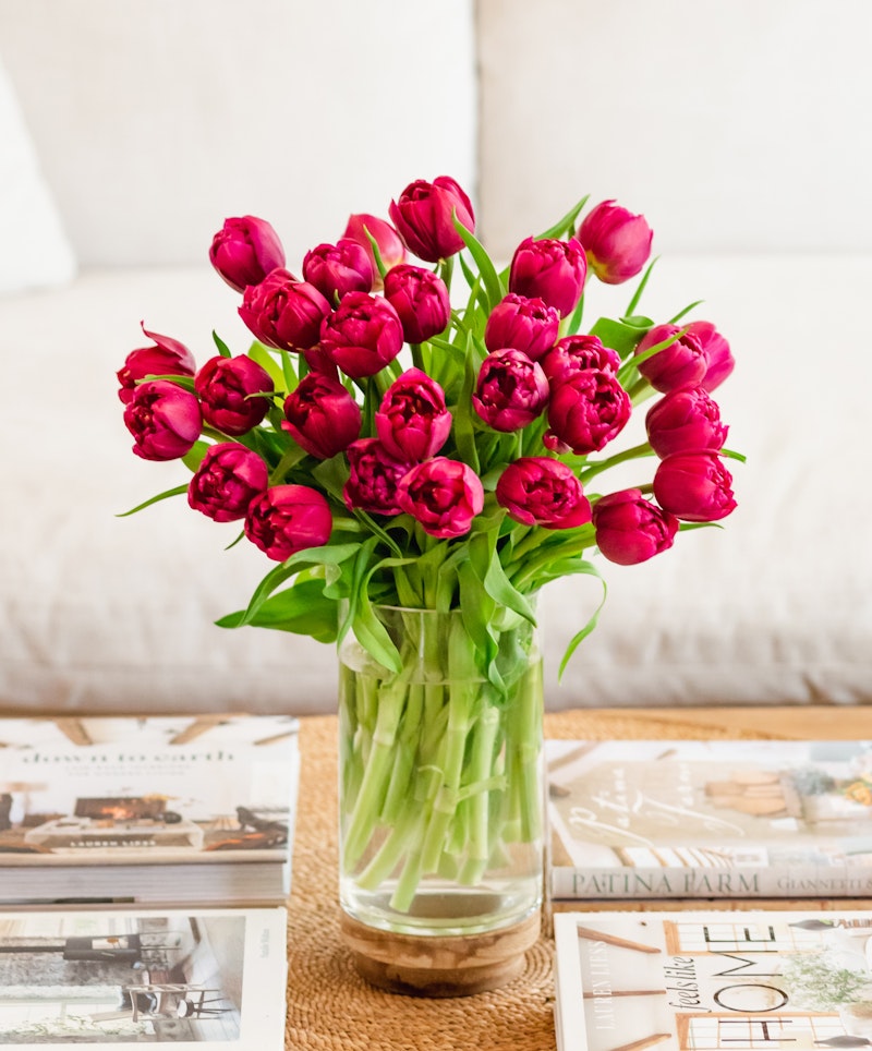 Vibrant bunch of pink tulips displayed in a clear vase on a wooden coffee table, with assorted magazines scattered around, creating a cozy home atmosphere.