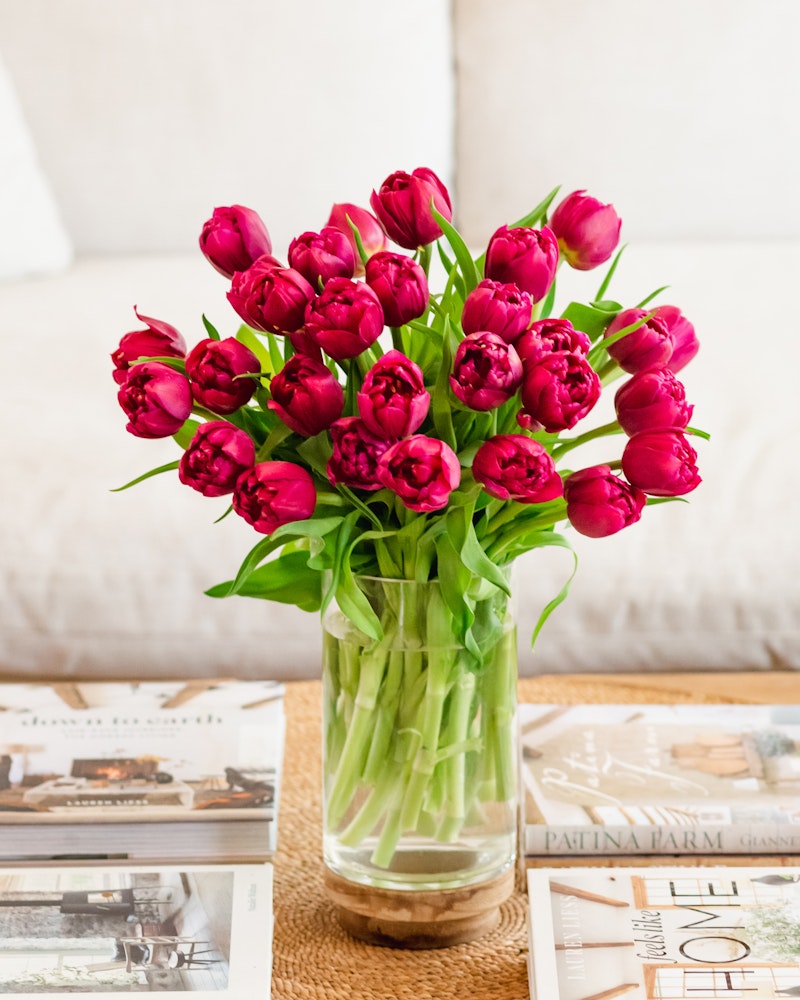 Vibrant bunch of pink tulips displayed in a clear vase on a wooden coffee table, with assorted magazines scattered around, creating a cozy home atmosphere.