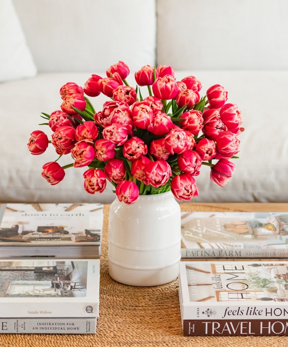 A vibrant bouquet of pink tulips in a white vase on a wooden table, surrounded by a collection of home decor magazines, creating a cozy and welcoming atmosphere.