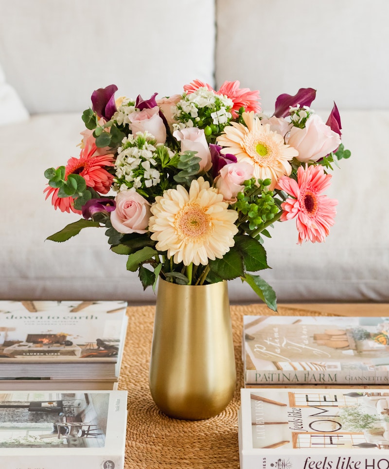 A vibrant bouquet of pink gerberas, purple tulips, white flowers, and greenery in a gold vase on a table with lifestyle magazines, creating a cozy home atmosphere.