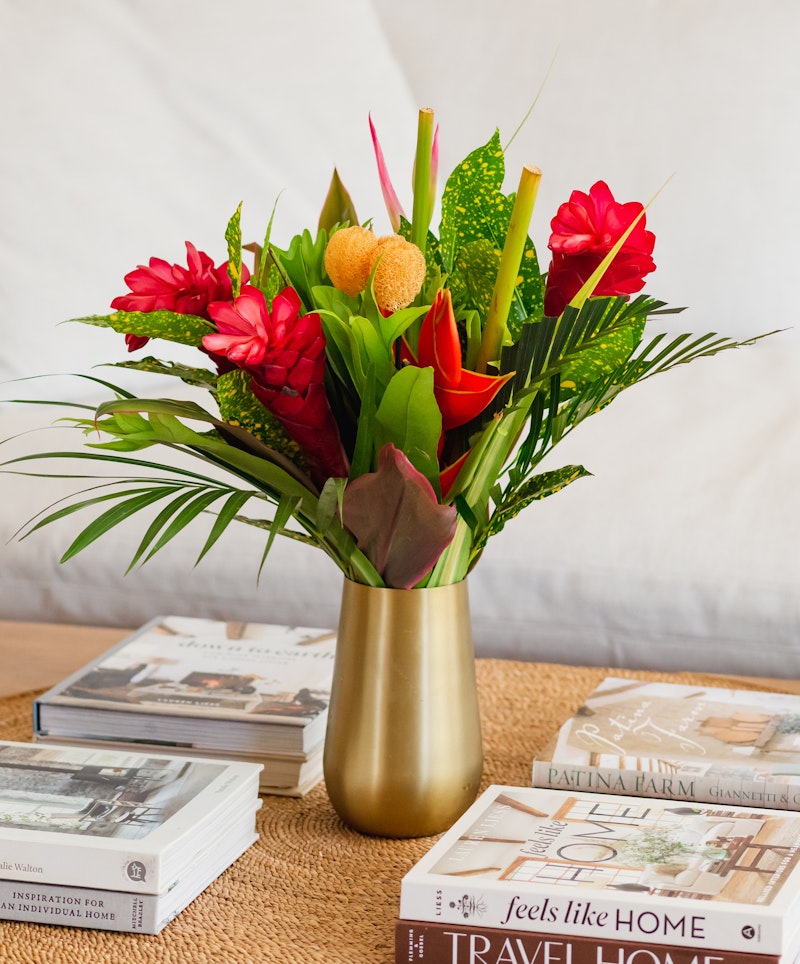 Vibrant tropical flower arrangement in a gold vase with red ginger flowers and lush green leaves, set atop coffee table books with a light backdrop.