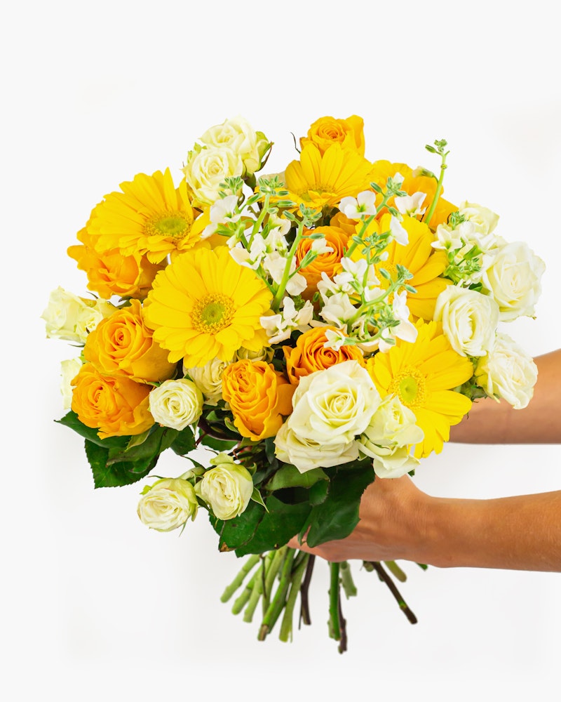 A vibrant bouquet of flowers held in a person's hands against a white background, featuring yellow blooms, orange roses, and delicate white accents.