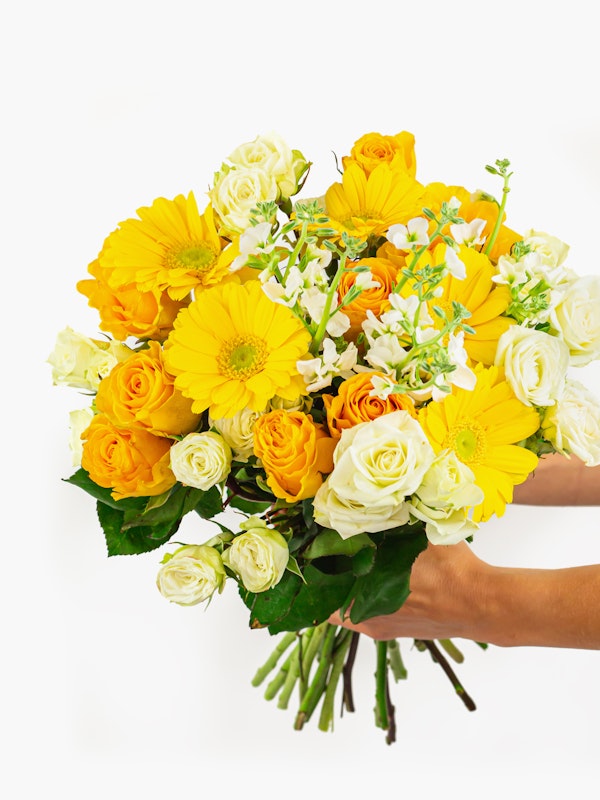 A vibrant bouquet of flowers held in a person's hands against a white background, featuring yellow blooms, orange roses, and delicate white accents.