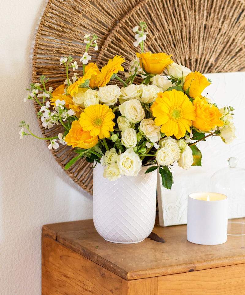Bright bouquet of yellow flowers and white roses in a white textured vase on a wooden table against a neutral wall with decorative woven plates.