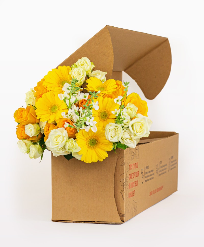 Bright bouquet of yellow and white flowers, including roses and gerberas, arranged in a cardboard gift box on a white background, suitable for various occasions.