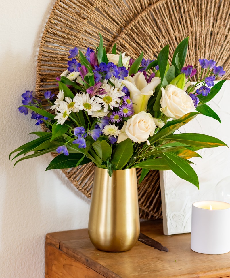 Bright bouquet of white roses, purple flowers, and green leaves in a golden vase on a wooden table, with woven round wall decor and a lit candle nearby.