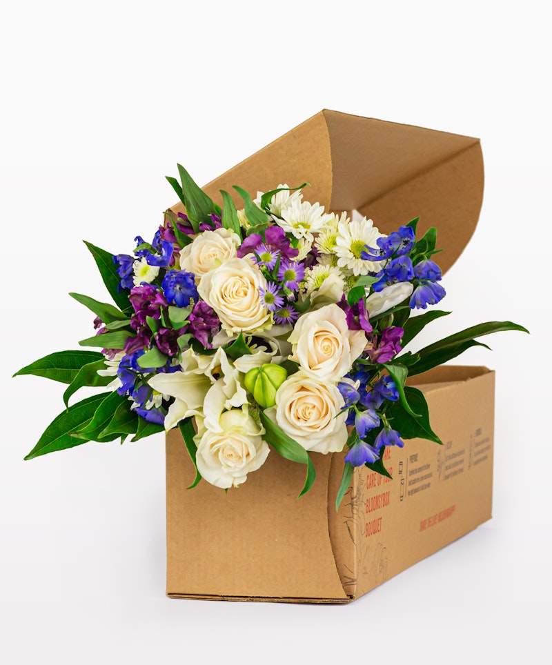 Vibrant bouquet of white roses and purple flowers emerging from a cardboard delivery box against a clear white background, showcasing fresh floral delivery service.