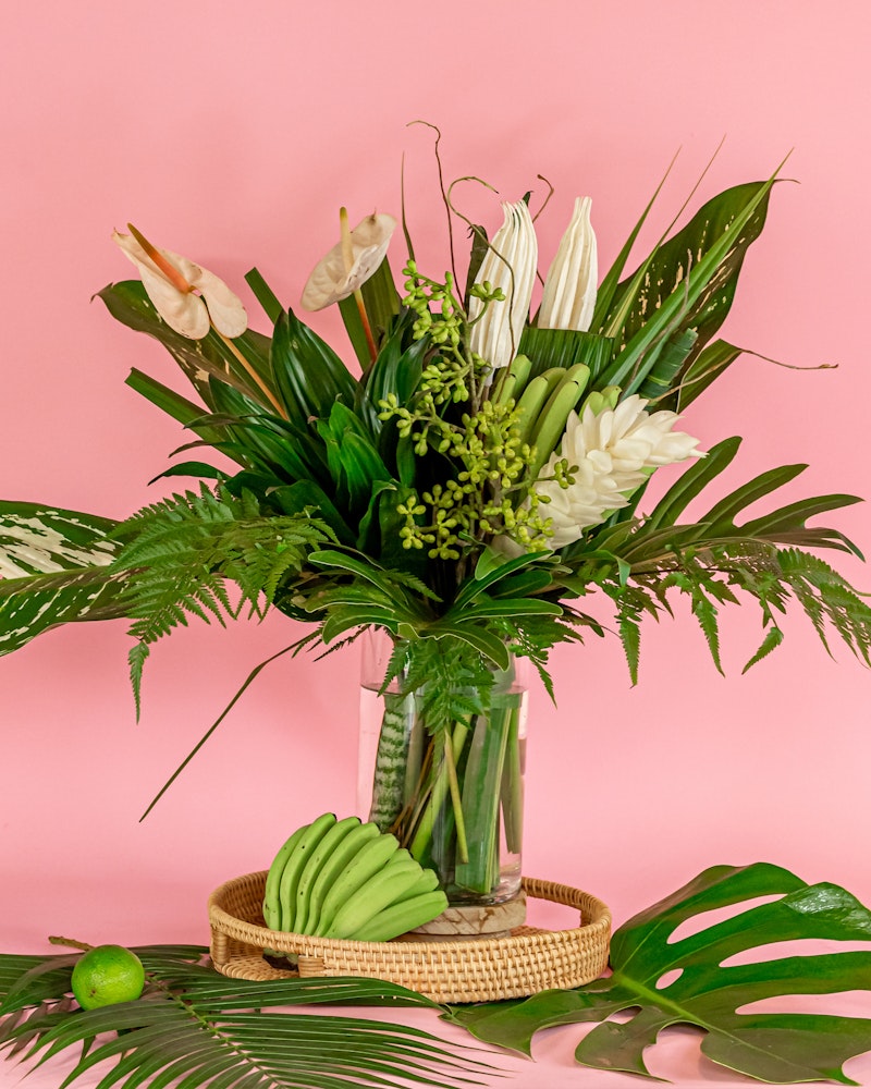 Elegant bouquet of tropical flowers and green foliage in a clear vase on a wicker tray, with a fan of green bananas and lime on a pink background.