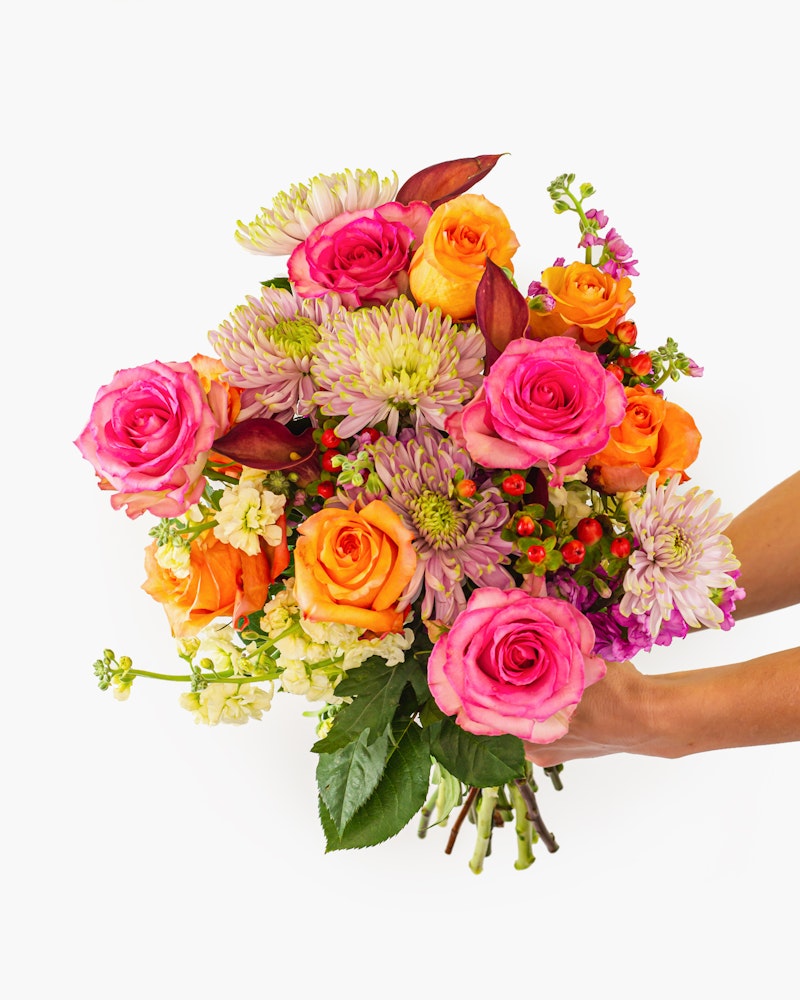 A vibrant bouquet of flowers held in a hand against a white background, featuring pink roses, orange blooms, and purple accents with green foliage.