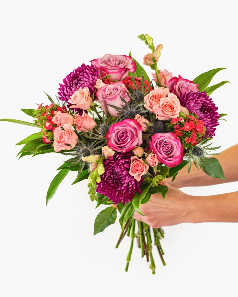 Vibrant bouquet of flowers including pink roses, purple chrysanthemums, and assorted greenery held in a hand against a white background, symbolizing a gift or celebration.