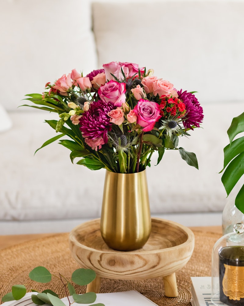 Elegant bouquet of pink roses and purple flowers arranged in a gold vase on a wooden table, with a cozy white sofa and green plant in the background.