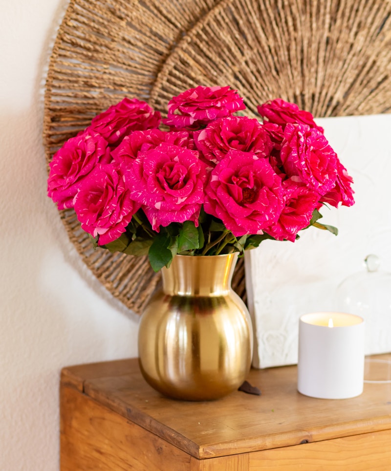 Vibrant pink roses arranged in a golden vase on a wooden table, complemented by a lit candle and a woven circular wall decor in a cozy room setting.