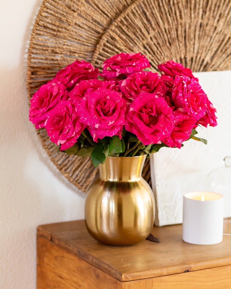 Vibrant pink roses arranged in a golden vase on a wooden table, complemented by a lit candle and a woven circular wall decor in a cozy room setting.