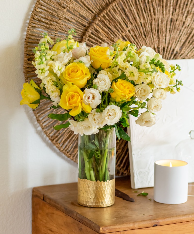 A vibrant bouquet of yellow roses and white blossoms in a clear vase with a golden base, placed on a wooden table beside a lit candle, with a woven decoration on the wall.