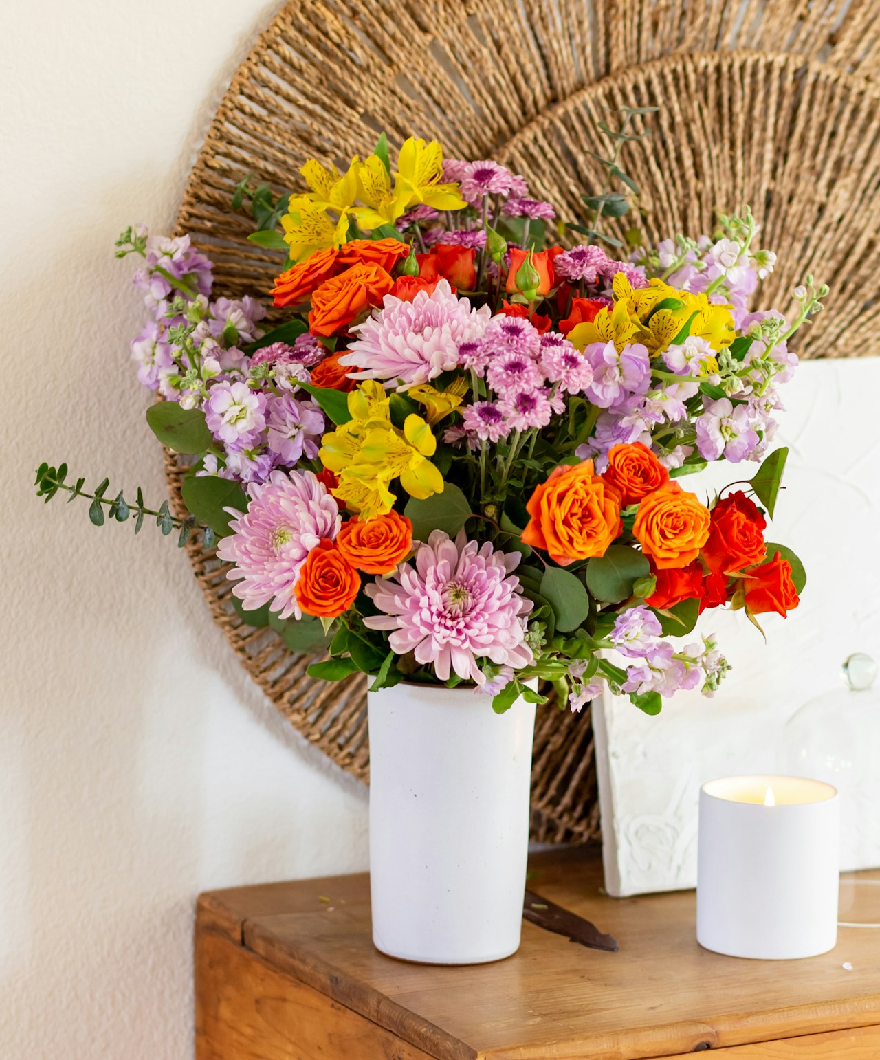Vibrant floral arrangement featuring orange roses, daisies, and lavender blooms in a white vase.